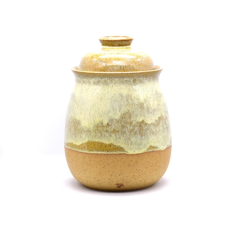 May include: A handmade ceramic jar with a lid. The jar has a rounded shape with a light brown base and a creamy, off-white upper section. The lid is a matching color. The jar is suitable for kitchen storage.