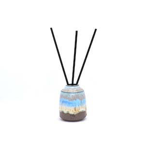 May include: A ceramic reed diffuser vessel with a rounded shape, featuring a reactive glaze in shades of light blue, cream, and brown. Three thin, straight black diffuser reeds are inserted into the narrow opening at the top of the container.