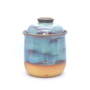 May include: A handmade ceramic jar with a lid. The jar is cylindrical with a light brown base and a turquoise and brown upper section. The lid is a matching turquoise and brown. The jar is likely used for storage.