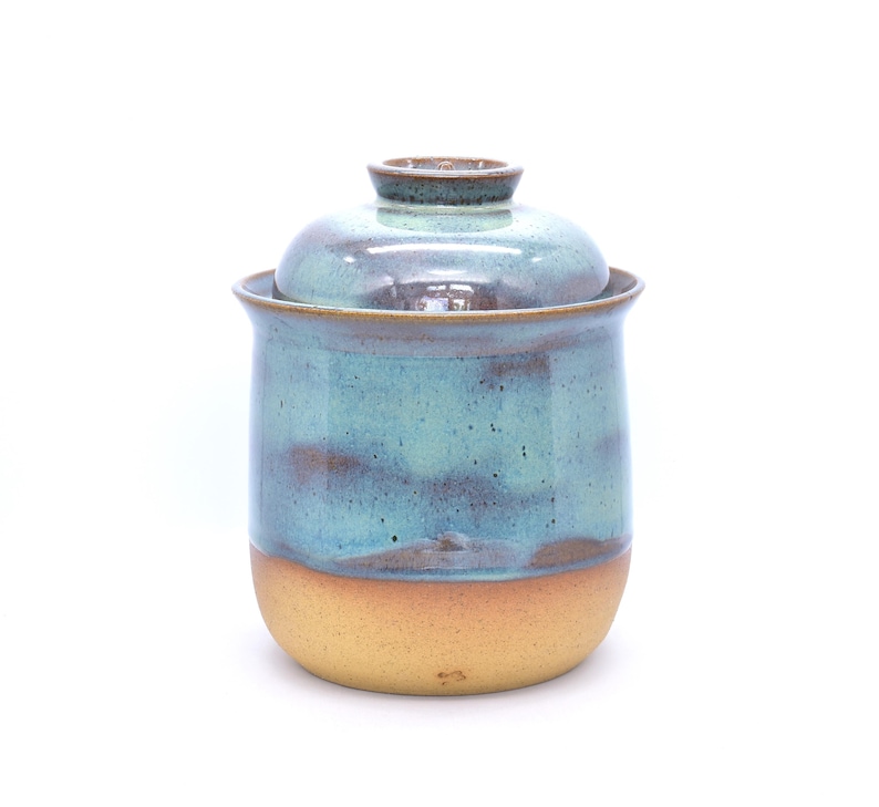 May include: A handmade ceramic jar with a lid. The jar is cylindrical with a light brown base and a turquoise and brown upper section. The lid is a matching turquoise and brown. The jar is likely used for storage.