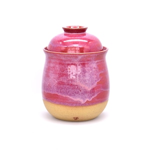 May include: A handmade ceramic jar with a lid. The jar is a vibrant pink with a lighter pink and cream-colored base. The jar has a rounded shape and a glossy finish, perfect for storing kitchen items.