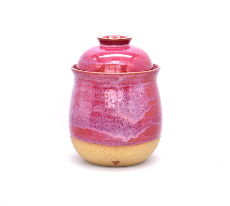 May include: A handmade ceramic jar with a lid. The jar is a vibrant pink with a lighter pink and cream-colored base. The jar has a rounded shape and a glossy finish, perfect for storing kitchen items.