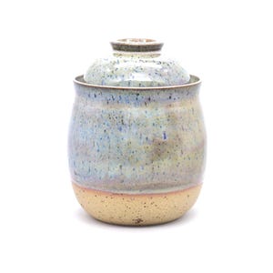 May include: A handmade ceramic jar with a lid. The jar has a rounded shape with a speckled, light blue and tan glaze. The base is a lighter tan color. The lid is also glazed in a similar pattern.