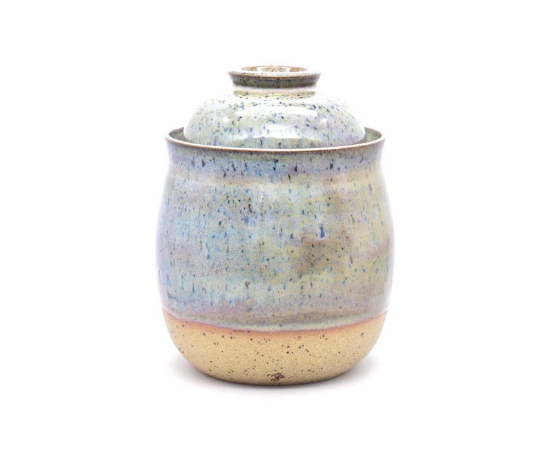 May include: A handmade ceramic jar with a lid. The jar has a rounded shape with a speckled, light blue and tan glaze. The base is a lighter tan color. The lid is also glazed in a similar pattern.