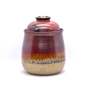 May include: A handmade ceramic jar with a lid. The jar is a deep red color with a gradient of brown and tan. The bottom portion is a speckled tan color. The lid is a matching red. This decorative jar is perfect for storing kitchen items.