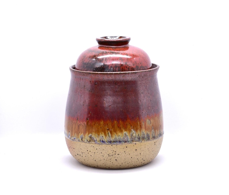May include: A handmade ceramic jar with a lid. The jar is a deep red color with a gradient of brown and tan. The bottom portion is a speckled tan color. The lid is a matching red. This decorative jar is perfect for storing kitchen items.