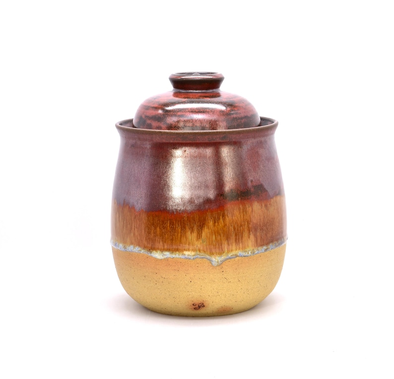 May include: A handcrafted ceramic jar with a lid. The jar has a rounded shape and is glazed in shades of red, brown, and yellow. The lid is a darker red. The jar is likely used for storage or decoration.