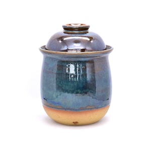 May include: A handmade ceramic jar with a lid. The jar has a rounded shape with a blue and teal glaze that transitions to a tan base. The lid is a darker brown color. The jar is likely used for storage or decoration.