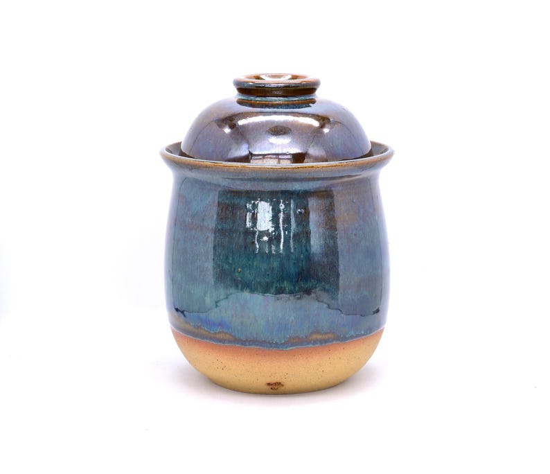 May include: A handmade ceramic jar with a lid. The jar has a rounded shape with a blue and teal glaze that transitions to a tan base. The lid is a darker brown color. The jar is likely used for storage or decoration.