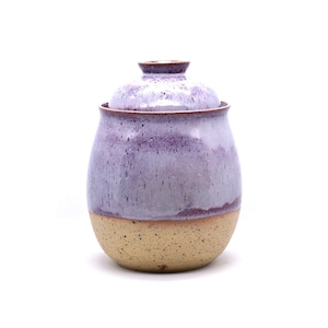 May include: A handmade ceramic jar with a lid. The jar has a rounded shape with a speckled lavender glaze on the upper portion and a tan, unglazed base. The lid is also lavender. The jar is likely used for storage.