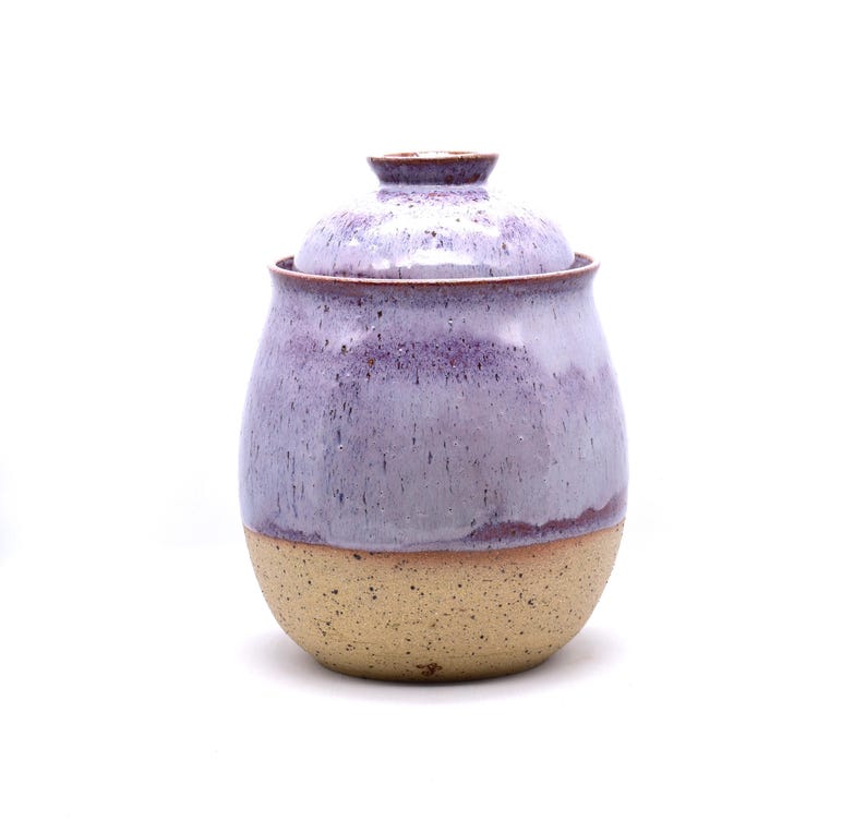 May include: A handmade ceramic jar with a lid. The jar has a rounded shape with a speckled lavender glaze on the upper portion and a tan, unglazed base. The lid is also lavender. The jar is likely used for storage.