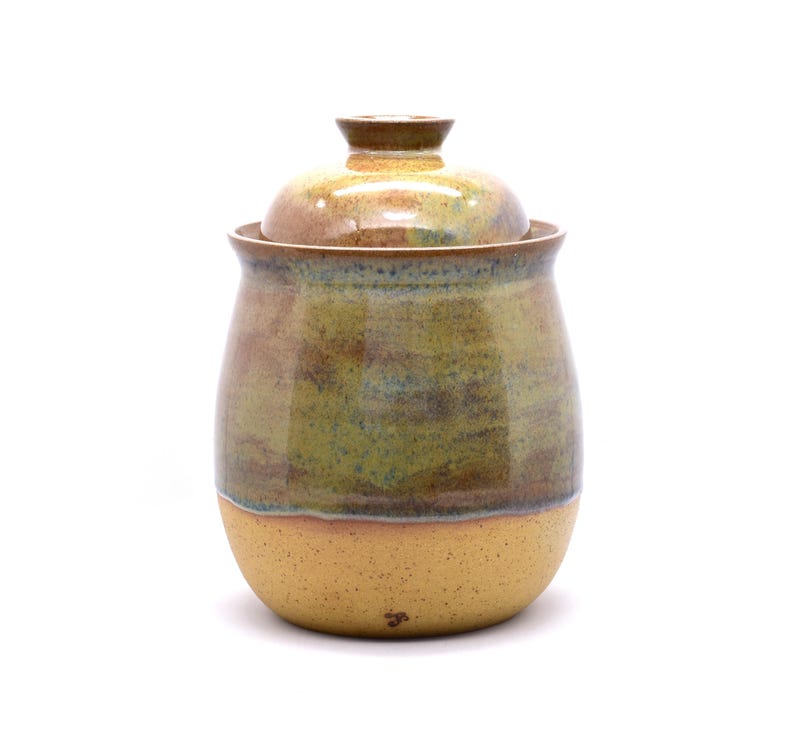 May include: A handcrafted ceramic jar with a lid. The jar has a rounded shape and is glazed in earthy tones of brown, green, and yellow. The lid sits atop the jar, completing the design. The jar is suitable for storing dry goods.