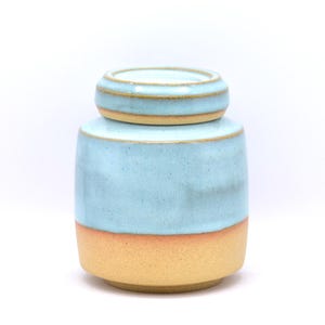 May include: A ceramic jar with a lid. The jar is light blue on top, transitioning to a tan base with an orange band. The lid is a smaller version of the jar, also light blue with a tan rim. The jar is likely used for storage.