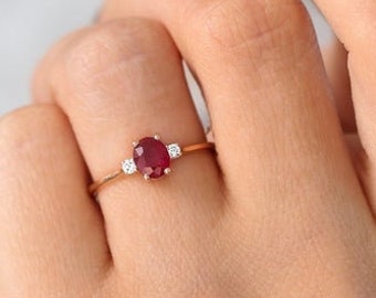 May include: A thin gold ring featuring a central oval-cut deep red ruby gemstone flanked by two small, round, clear white diamonds on either side. The band is slender and polished, showcasing a classic three-stone setting design.