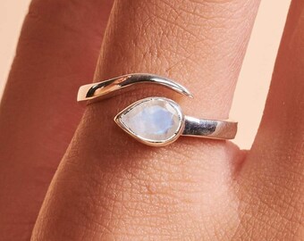 May include: A bypass ring crafted from polished silver, featuring a single pear-shaped moonstone set in a bezel. The band has a smooth, reflective finish and curves around the finger, with one end tapering to a point and the other holding the stone.