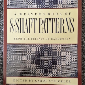 Op de afbeelding: Een paperback boek getiteld A Weaver's Book of 8-Shaft Patterns from the Friends of Handwoven, onder redactie van Carol Strickler. De kaft heeft een crèmekleurige achtergrond met donkerbruine randen en blauw-witte geweven patronen.