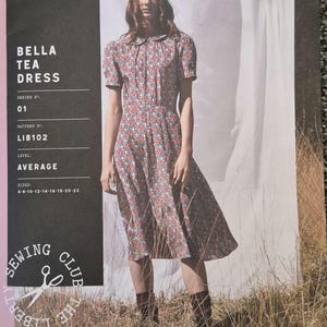May include: A woman wearing a floral print Bella Tea Dress. The dress has short sleeves and a Peter Pan collar. The image also shows the pattern details, including the Liberty Fabrics logo, series number, pattern number, level, and sizes.