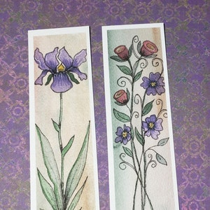 May include: Two watercolor bookmarks featuring floral designs. One bookmark displays a purple iris with green leaves. The other shows red roses and purple flowers with green leaves and stems. Both are on a white background.