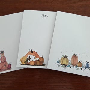 May include: Three white rectangular notepads featuring watercolor illustrations of pumpkins. The left pad shows a stack of pumpkins, the center pad displays a goose with pumpkins and the text Notes, and the right pad features a row of pumpkins.
