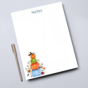 May include: A white notepad with the word Notes printed in blue at the top. The bottom left corner features a watercolor illustration of a stack of four pumpkins in orange, green, and blue, surrounded by small leaves and flowers. A beige pen.