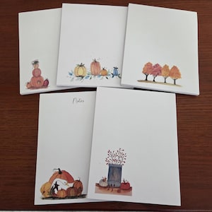 May include: Five rectangular white paper notepads featuring autumn-themed watercolor illustrations. Designs include stacked pumpkins, a pumpkin patch, a row of fall trees, a goose with pumpkins, and a jar with branches and pumpkins. One pad says Notes.