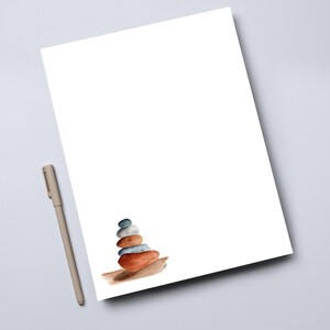 May include: A white sheet of paper with a watercolor painting of a stack of stones in shades of blue, gray, and orange. A beige pen rests on the gray surface next to the paper.