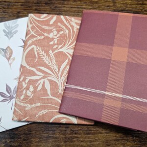 May include: Three rectangular notebooks with distinct covers. The first features a white background with scattered brown and gray autumn leaves. The second has a terracotta background with a white wheat stalk pattern. The third is a dark red plaid.