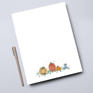 May include: A white sheet of paper with a watercolor illustration of four pumpkins in shades of orange, yellow, and blue, with green leaves. A beige pen rests on the left side of the paper. The pumpkins are arranged in a row.