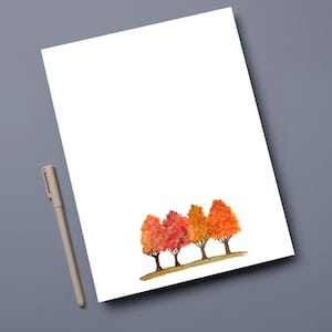 May include: White stationery paper with a watercolor painting of four trees in autumn colors. The trees are shades of red and orange. A beige pen rests on a gray surface next to the paper.