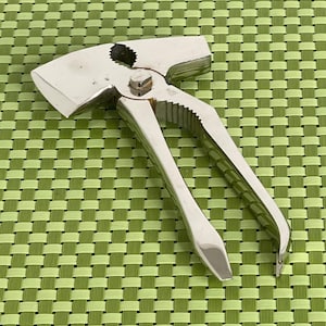 May include: A shiny, silver nutcracker with a lever handle and a toothed cracking surface. The nutcracker has a hammer-like head and is designed for cracking nuts. It is shown on a green woven placemat.