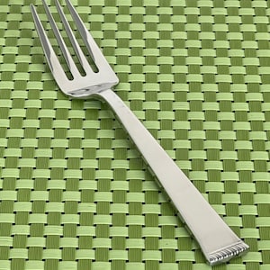 May include: A stainless steel fork with four tines and a rectangular handle. The handle has a decorative edge. The fork is set against a green woven placemat.