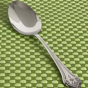 May include: A silver spoon with a floral design on the handle. The spoon is on a green woven placemat.