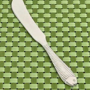 May include: A stainless steel butter knife with a fish-shaped handle. The blade is smooth and rectangular, while the handle features a textured fish scale design and a fish head at the end. The knife is set against a green woven background.