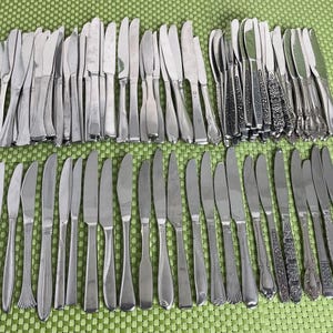 May include: A large collection of assorted stainless steel dinner knives arranged in two rows. The set includes various handle designs, ranging from plain, smooth surfaces to intricate embossed patterns, all featuring serrated or straight blades.