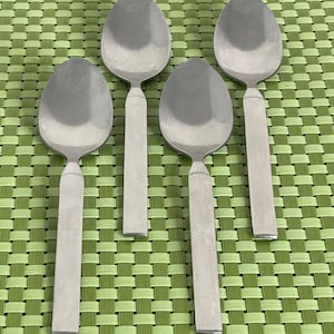 May include: Four silver-colored metal spoons arranged in two rows. Each spoon features a smooth, oval-shaped bowl and a flat, rectangular handle that tapers slightly where it meets the bowl. The metal has a consistent, polished finish.
