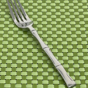 Peut inclure: Une fourchette en acier inoxydable à quatre dents et un manche de style bambou. La fourchette est posée sur un fond tissé vert et vert clair, créant un contraste de textures et de couleurs. La fourchette est conçue pour les repas.