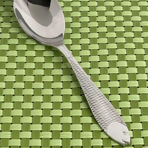 May include: A silver spoon with a fish design on the handle. The spoon is on a green woven placemat.