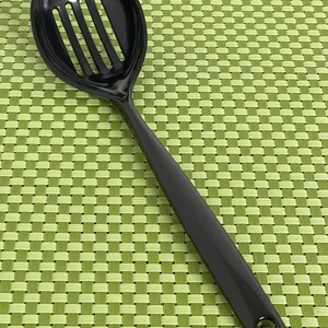 Peut inclure: Une louche noire à fentes avec un long manche et un trou pour suspendre. La tête de la cuillère est arrondie avec des fentes verticales. La cuillère est en matériau brillant, sur un fond vert tissé.