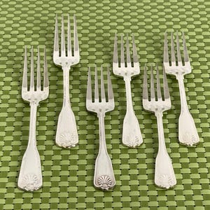 May include: Seven silver forks with ornate handles and tines are arranged on a green patterned surface. The forks have a decorative shell design at the base of the handle. The tines are evenly spaced.