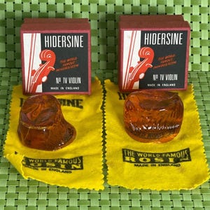 May include: Two red boxes with the text "HIDERSINE" and "THE WORLD FAMOUS IMPROVED RESIN NO IV VIOLIN MADE IN ENGLAND" on them. The boxes are sitting on yellow cloths with the text "THE WORLD FAMOUS ROSIN MADE IN ENGLAND" on them. There are two amber colored violin rosin cakes on the cloths.