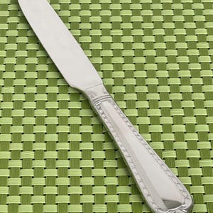 May include: A silver butter knife with a decorative pattern on the handle. The knife is on a green woven placemat.