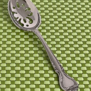 May include: A silver-toned metal slotted serving spoon featuring a polished finish, an oval bowl with a decorative cut-out pattern, and a handle adorned with embossed scrollwork details at the neck and a floral motif at the base.