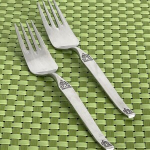 May include: Two silver forks with ornate designs on the handles. The forks are on a green woven placemat.