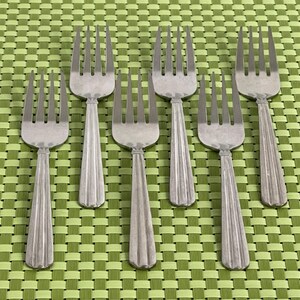 May include: A set of six silver-toned metal forks arranged in two rows. Each fork features four tines and a handle with three vertical ridges running down the center. The forks have a polished finish and a uniform design with a rounded neck.