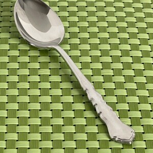 May include: A silver spoon with a textured handle. The spoon is on a green woven placemat.
