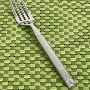 May include: A stainless steel fork with a light gray handle featuring three small white dots. The fork has four tines and a polished finish. The handle has a rectangular end. The fork is on a green woven placemat.