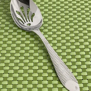 May include: A silver slotted spoon with a fish design on the handle. The spoon is on a green woven placemat.