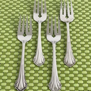 May include: Four silver forks with ornate handles are arranged on a green woven placemat. The forks have three tines and decorative detailing at the base of the handle. The handles have a floral design at the base.