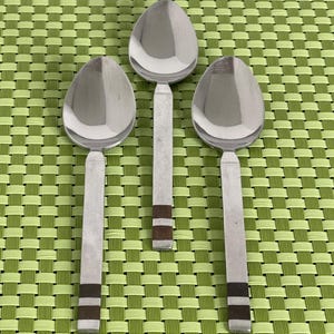 May include: Three silver spoons with black and brown stripes on the handles. The spoons are arranged in a row on a green woven placemat.