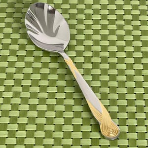 May include: A stainless steel serving spoon with a polished finish. The handle features a gold-colored, decorative design. The spoon is set against a green woven placemat.
